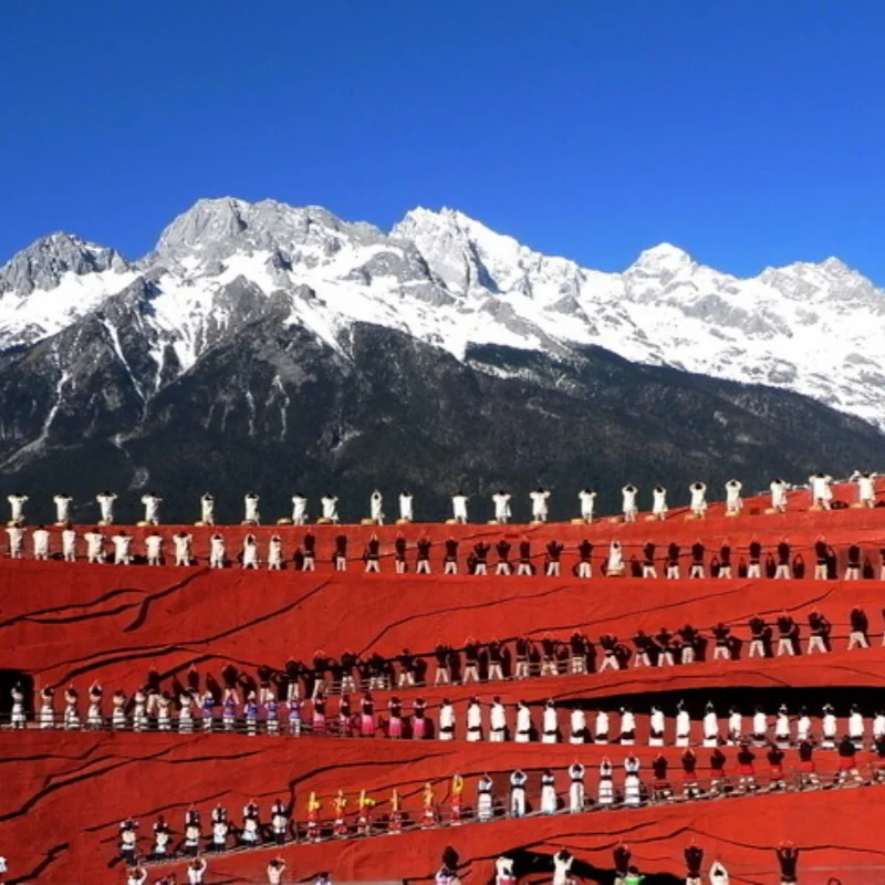 Impression Lijiang Performance 1 印象麗江大型實景表演在玉龍雪山下上演,紅色階梯舞台上眾多演員身著民族服飾整齊排列演出,背後是壯麗的雪山與藍天景觀,展現麗江獨特的民族文化與印象麗江震撼的戶外表演場面