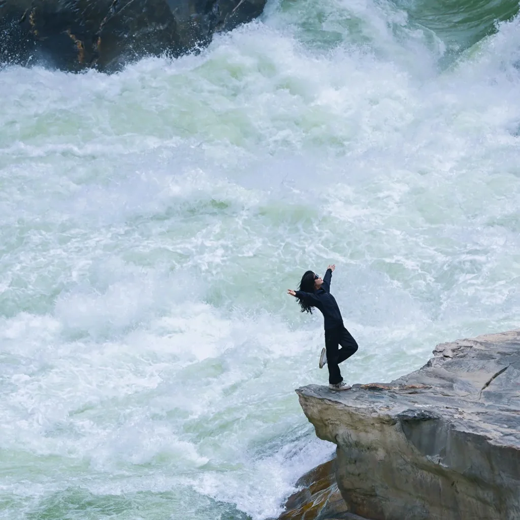 Lower Tiger Leaping Gorge Viewing Platform 在 虎跳峽 下段觀景台,一名遊客站在岩石邊緣張開雙臂,腳下江水奔騰翻湧,激起層層白浪,場面震撼壯觀。