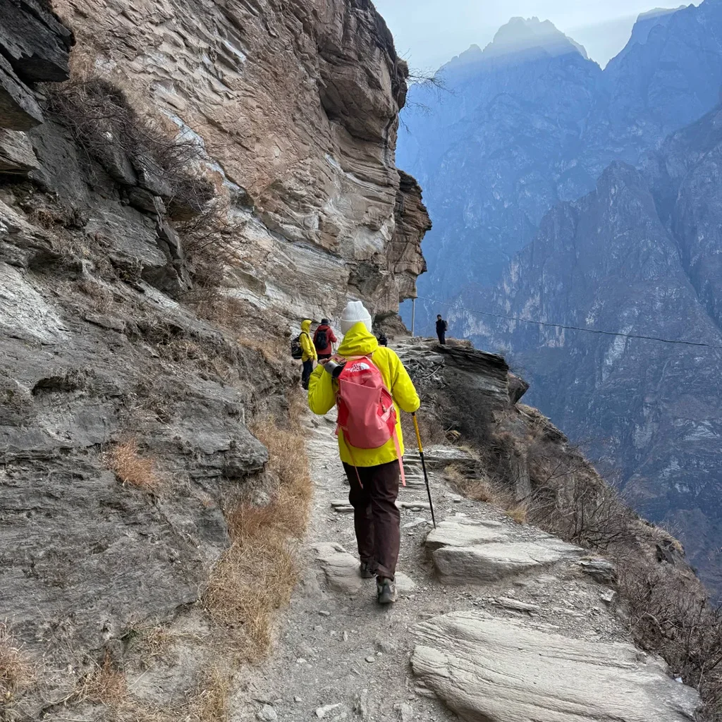 Middle Tiger Leaping Gorge Hiking 在 虎跳峡 悬崖栈道上徒步的游客,背着登山包沿着崎岖山路前行,峡谷深邃壮观,远处山峰连绵,展现惊险与壮美并存的自然风光。