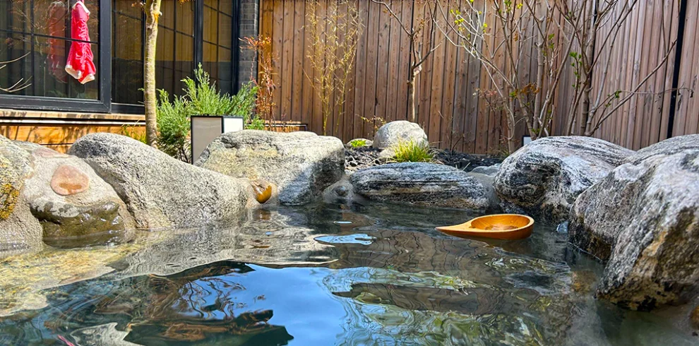 SPA 騰衝溫泉庭園露天湯池景觀,清澈溫泉水環繞天然岩石與日式庭園造景,木牆與綠樹點綴寧靜環境,木製湯勺漂浮水面,呈現騰衝溫泉放鬆療癒的泡湯氛圍,是雲南著名的溫泉養生旅遊景點。