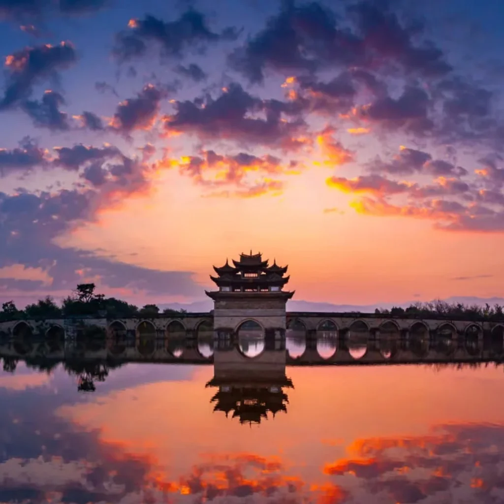 Shuanglong Bridge 建水雙龍橋夕陽景觀,古老石拱橋橫跨水面,橋中央的傳統亭閣在晚霞映照下形成優美剪影,橋洞與天空雲彩倒映在平靜水面上,呈現雙龍橋典雅壯麗且充滿歷史韻味的古橋風光