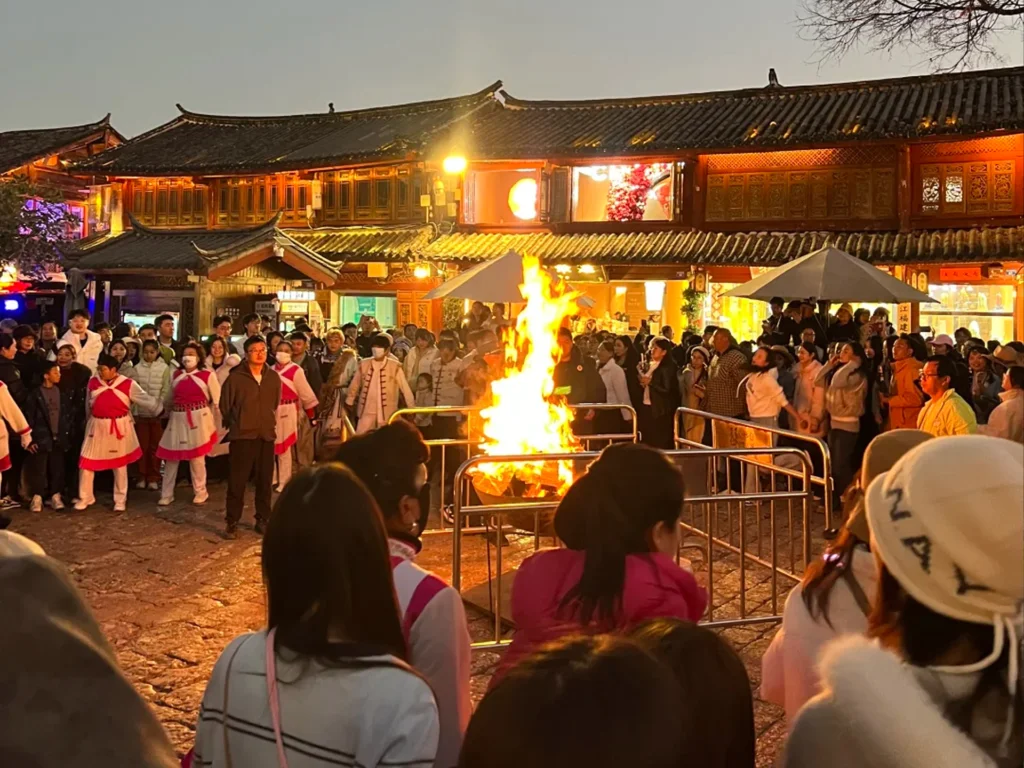 雲南麗江古城四方街篝火晚會，群眾圍繞熊熊篝火，背景是傳統納西建築與暮色天空。