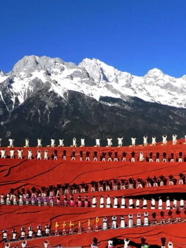 Impression Lijiang Performance 1 印象麗江大型實景表演在玉龍雪山下上演,紅色階梯舞台上眾多演員身著民族服飾整齊排列演出,背後是壯麗的雪山與藍天景觀,展現麗江獨特的民族文化與印象麗江震撼的戶外表演場面