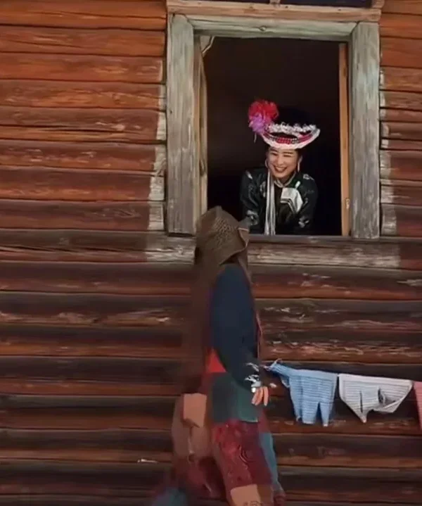 Mosuo family visit1 一位身著傳統服飾的摩梭女子從木楞房窗戶探頭微笑,與窗外的訪客互動,展示摩梭族獨特的建築與人文風情。