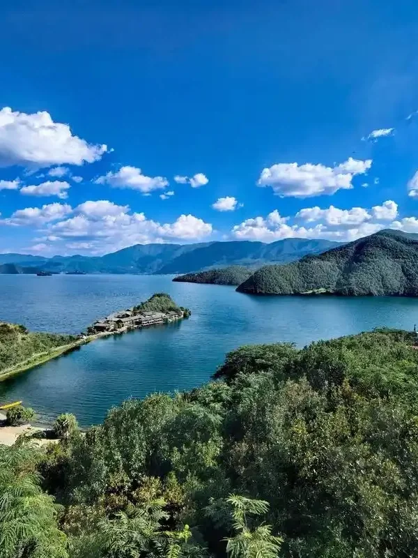 Panoramic view of Lugu Lake 瀘沽湖壯麗湖景在晴朗藍天下展開,湖水深藍清澈,群山環繞湖泊。湖中央的小島與連接岸邊的堤道清晰可見,周圍綠意盎然的森林與遠方山巒層層延伸,白雲點綴天空,展現瀘沽湖高原湖泊寧靜而壯觀的自然風光。